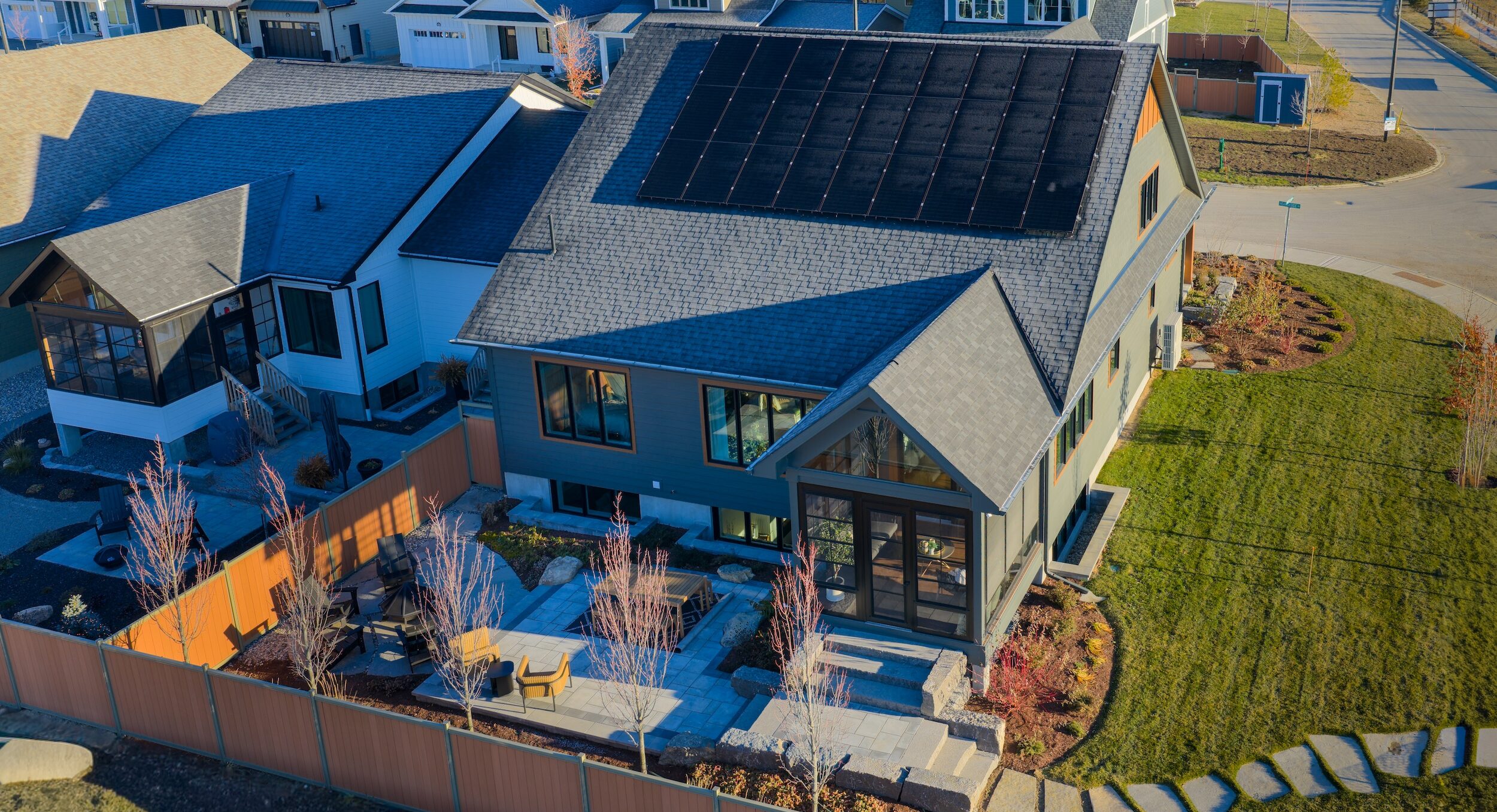 A modern net zero house with solar panels on the roof, a landscaped backyard with stone paths and patio seating, surrounded by a wooden fence in a suburban neighborhood.