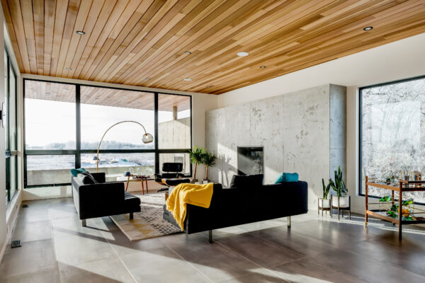 Modern living room with large windows, wooden ceiling, two black sofas, a yellow blanket, indoor plants, and a cozy fireplace. Natural light fills the space, highlighting the minimalist decor and scenic outdoor view—perfect inspiration for those seeking renovation ideas or exploring Home Renovation trends recognized by the CHBA National Awards for Housing Excellence.