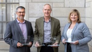 Three adults in business attire stand side by side, each holding a glass award. They are smiling, posing in front of a light-colored stone wall with a window on the left side.