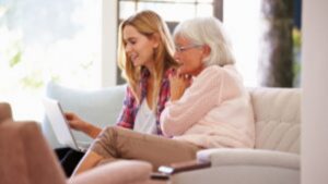 A young woman and an older woman sit together on a couch, smiling and looking at a laptop screen in a bright, comfortable living room.
