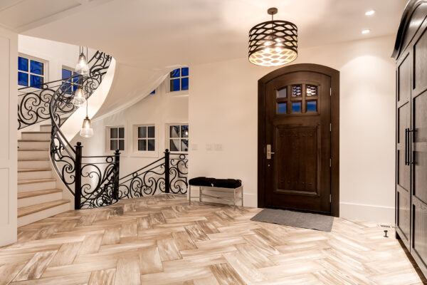 Modern foyer with wood herringbone floor, dark wooden front door, black cushioned bench, ornate curved staircase with decorative railing, pendant lights, and white walls—showcasing fabulous flooring that inspires fresh renovation ideas for your next home renovation project.