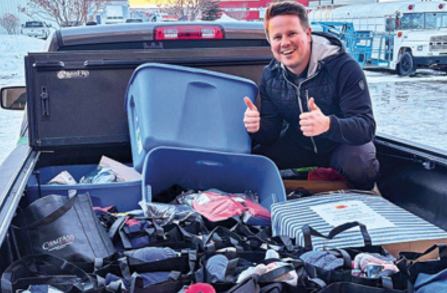 A smiling man gives two thumbs up while standing next to a truck bed full of bags, bins, and boxes filled with assorted items, outside in a snowy area.