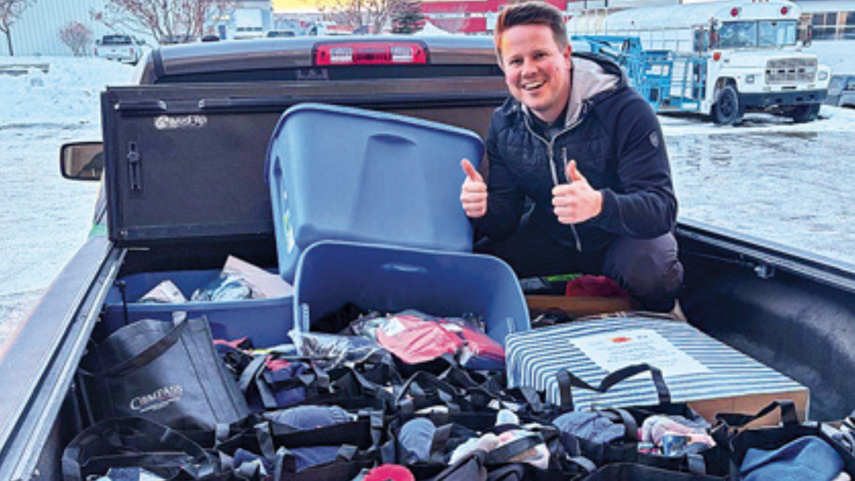 A smiling man gives two thumbs up while standing next to a truck bed full of bags, bins, and boxes filled with assorted items, outside in a snowy area.