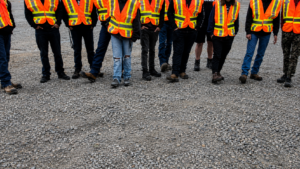 A group of people wearing orange and yellow safety vests from CHBA Fraser Valley stand on gravel, shown from the shoulders down with their faces out of frame—perhaps gathered for a boot bank initiative in the Fraser Valley.