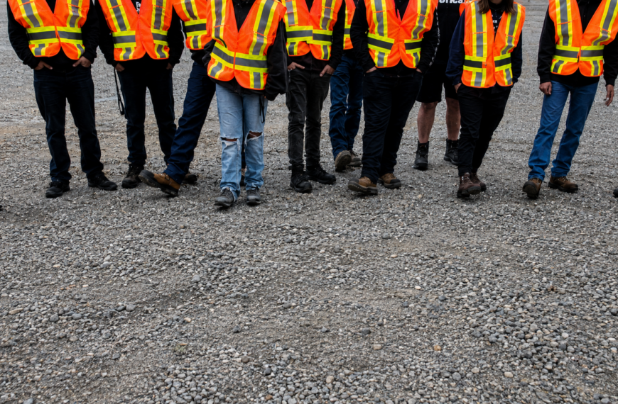A group of people wearing orange and yellow safety vests from CHBA Fraser Valley stand on gravel, shown from the shoulders down with their faces out of frame—perhaps gathered for a boot bank initiative in the Fraser Valley.
