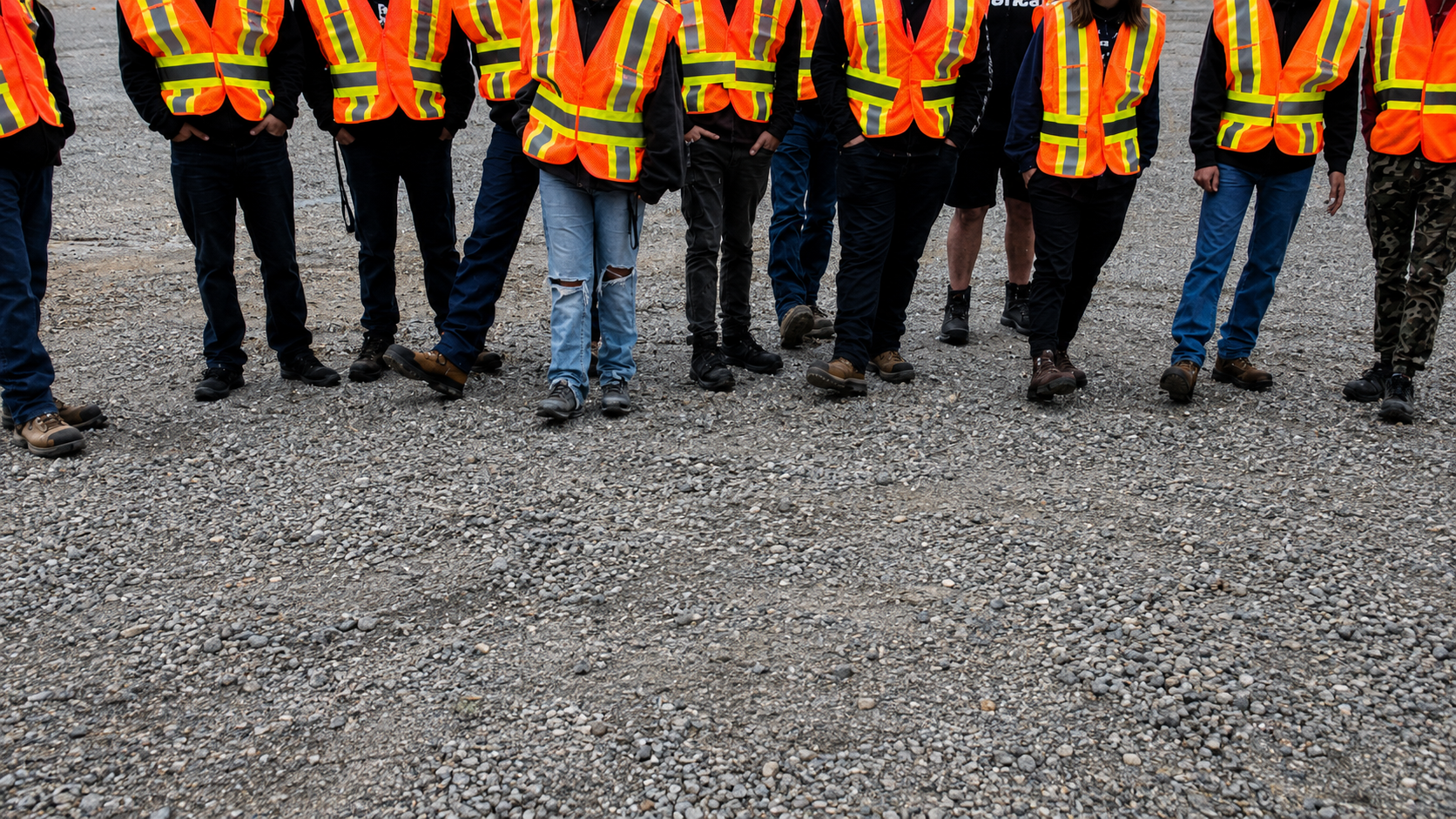A group of people wearing orange and yellow safety vests from CHBA Fraser Valley stand on gravel, shown from the shoulders down with their faces out of frame—perhaps gathered for a boot bank initiative in the Fraser Valley.