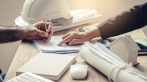 Two people sit at a desk with documents, one signing papers. Nearby are a small wooden house model, a hard hat, blueprints, keyboard, and a computer mouse, suggesting a construction or real estate meeting. The scene could easily illustrate a guide to renovation contracts, as it captures the essential elements of the planning and agreement process in property development or remodeling projects.