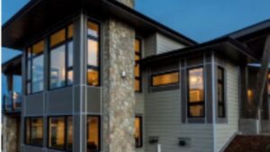 Two-story modern house with large windows, stone chimney, and horizontal siding, photographed at dusk with interior lights on.