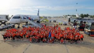 A large group of people in matching orange shirts poses on an airport tarmac near an airplane and several buses under a partly cloudy sky.