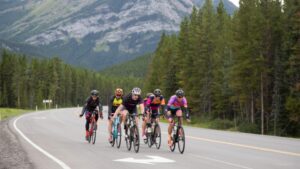 Five cyclists ride on a winding mountain road surrounded by dense green trees and tall rocky mountains under an overcast sky.