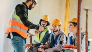 A construction supervisor explains plans to three workers wearing yellow helmets and safety vests. Two workers take notes while listening attentively inside a partially finished building.