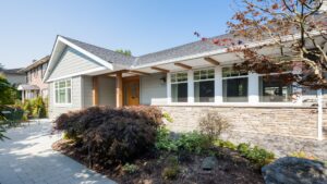 Single-story house with gray siding, large windows, and a stone accent wall. A landscaped garden with shrubs, small trees, and a walkway leads to the front door. Sunny, clear weather.