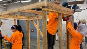 Four people in bright orange shirts work together building a wooden structure indoors at the Jill of All Trades event, with participants from North Okanagan high schools. One person drills into a beam while others assist, surrounded by workshop tools and equipment.