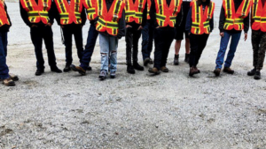 A group of people wearing orange high-visibility safety vests and standing on a gravel surface. Their faces are out of frame; only their legs and torsos are visible.