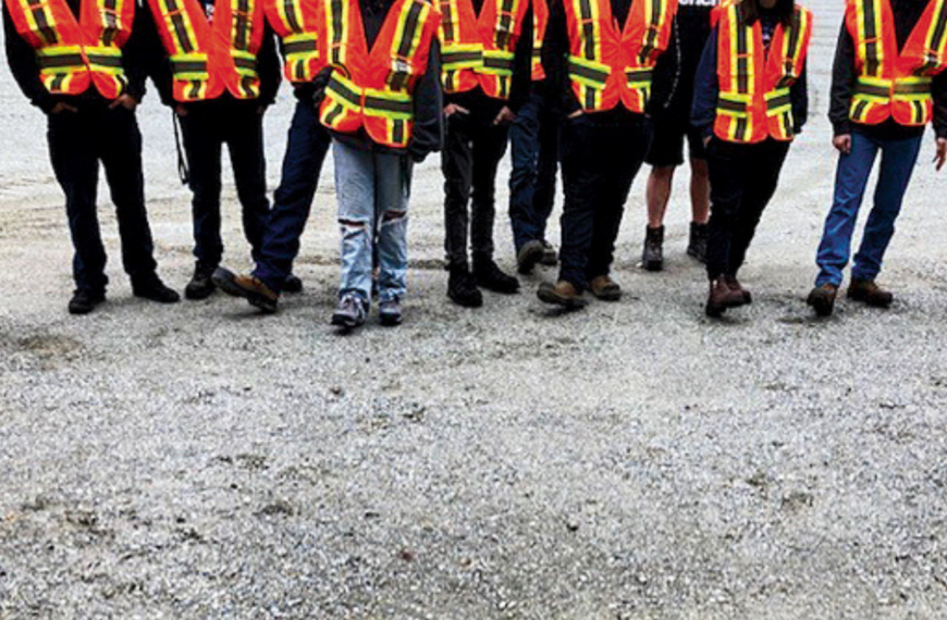 A group of people wearing orange high-visibility safety vests and standing on a gravel surface. Their faces are out of frame; only their legs and torsos are visible.