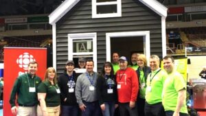 A group of people stand smiling in front of a small dark-gray house with white trim indoors; some wear bright green shirts, others wear green, red, or dark tops. A CBC banner stands to the left.