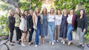A group of eleven women stand closely together outdoors, smiling at the camera. They are casually dressed and standing on a pathway surrounded by greenery and flowers.