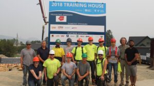A group of construction workers in hard hats pose in front of a large 2018 Training House sponsor sign at a building site. The workers are standing and kneeling on a wooden platform.