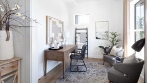 Bright home office with a wooden desk, black chair, lamp, framed art, ladder shelf with blankets, plants, and two gray armchairs on a patterned rug. Large window lets in natural light.