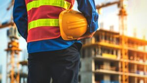 A construction worker wearing a reflective vest holds a yellow hard hat while facing a building under construction, with cranes and scaffolding visible in the background.