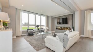 Modern living room with large windows, white sofa, gray rug, glass coffee table, two black chairs, wall-mounted TV above a fireplace, and neutral-toned decor. Natural light fills the room.