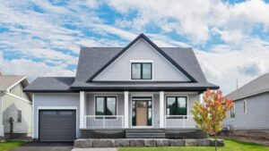 A modern gray house with white trim, a front porch, single-car garage, and a small tree in the yard, set against a partly cloudy blue sky.