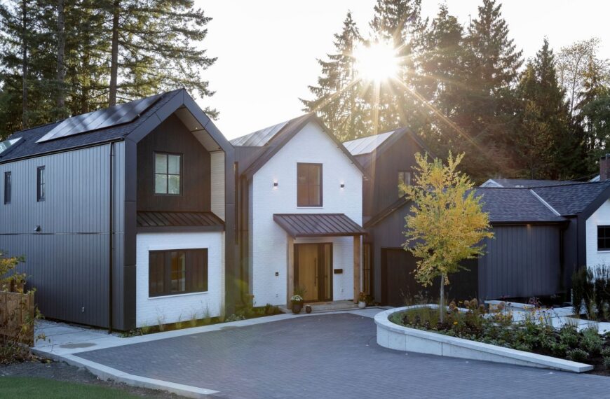 Modern two-story house with dark metal and white brick exterior, landscaped front yard, driveway, and sunlight shining through tall trees in the background.