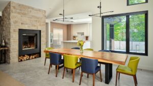 Modern dining room with a wooden table, blue and green chairs, a vase of flowers, large windows, stone fireplace with firewood, and a bright kitchen area in the background.