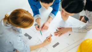 Three people view and discuss architectural blueprints spread out on a table. Two point with pencils, and one looks on thoughtfully. A yellow hard hat and ruler lie nearby. The image is shot from above.