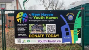 A banner on a fence reads A New Haven for Youth Haven: Building Brighter Futures Together! with a QR code, donation thermometer, home photo, and a call to donate at YouthHaven.ca to support youth shelter funding.