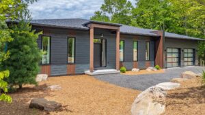 A modern single-story house with dark wood and vertical brown accents, large windows, a black front door, and a double garage, surrounded by mulch, rocks, and green trees.