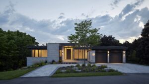 Modern, single-story house with large windows, stone and wood exterior, two-car garage, landscaped front yard, and a tree, photographed at dusk with lights glowing inside.