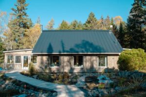 A modern wooden house with a dark metal roof sits among trees and a garden. A curved stone path leads to the entrance, and the sky is clear and blue.