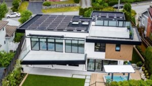 A modern two-story house with solar panels on the roof, large glass windows, a patio area, green lawn, and a covered outdoor hot tub, situated in a suburban neighborhood.