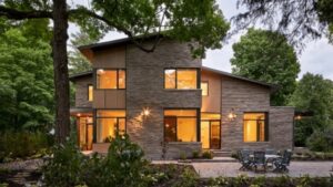 Modern two-story house with large windows, stone exterior, and warm interior lights, surrounded by trees and greenery. Outdoor patio with a table and chairs is visible in the foreground.