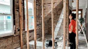 A person in a red shirt stands in a room under renovation, looking at exposed wall studs and lath. A ladder and construction debris are visible, with light coming in through nearby windows.