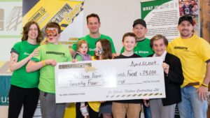 A group of people, some in yellow shirts and face paint, hold a large $24,000 check for The Isaac Foundation Research Fund, dated April 22, 2018. Smiling children and adults pose in front of informational banners.