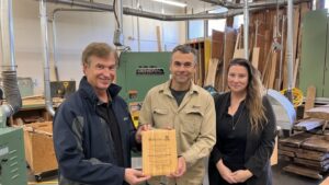 Three people stand in a woodworking shop, two men and one woman. The man on the left holds a wooden plaque with engraved text. Woodworking equipment and wooden boards are visible in the background.