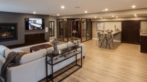Modern living room with a fireplace, wall-mounted TV, light wood floors, an L-shaped sofa, bar area with marble countertop, and a glass-enclosed wine cellar. Neutral colors and recessed lighting create a cozy, upscale atmosphere.