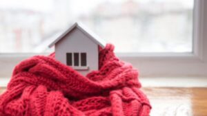 A small model house wrapped in a thick red knitted scarf sits on a windowsill, symbolizing warmth and home heating.