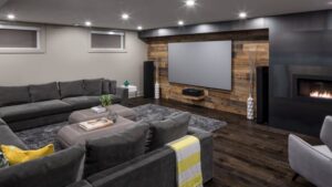 A modern home theater room with a large gray sectional sofa, a projector screen on a wood accent wall, a fireplace, speakers, a gray area rug, and dark wood floors. Decorative items are on the ottoman and shelves.