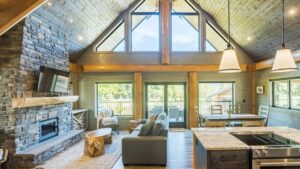 Spacious cabin living room with high vaulted ceiling, large windows, stone fireplace, gray sofa, rustic wood furniture, and an open kitchen with island lighting, overlooking a sunny outdoor deck.
