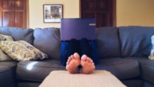 A person sits on a gray couch with bare feet up on an ottoman, using a laptop that obscures their face. Pillows and framed art are visible in the background.