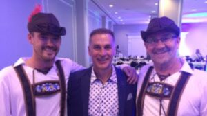 Three men pose together at an indoor event connected to the Street Culture Project; two wear traditional Bavarian outfits with hats and suspenders, while the man in the center dons a suit jacket and a polka-dot shirt.