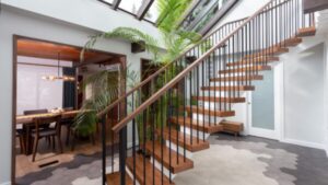 Modern open-plan interior with a floating wooden staircase, black railings, skylight, large indoor plant, hexagon tile floor, and a dining area visible through a wide doorway.