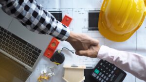 Two people shake hands over blueprints, with a laptop, calculator, tape measure, hard hat, and tools on the table, suggesting a construction or engineering agreement.