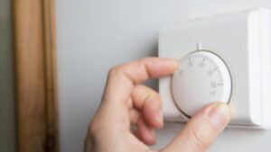 A hand adjusts a round white thermostat mounted on a wall, turning the dial to set the temperature.
