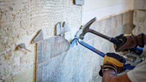 A person wearing work gloves uses a hammer and crowbar to remove ceramic tiles from a wall during a renovation project.