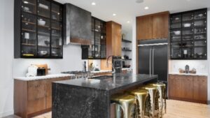 Modern kitchen with black countertops, wooden cabinets, glass-front uppers, a large island with gold stools, stainless steel appliances, and under-cabinet lighting. The space is bright and neatly organized.