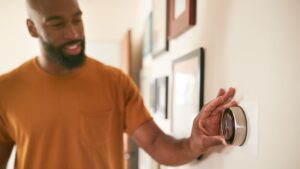 A man in an orange shirt adjusts a smart thermostat on a wall, surrounded by framed pictures.